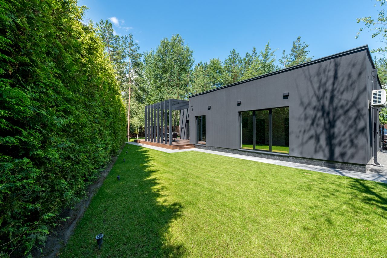 Sleek black modern house with large windows and vibrant green lawn surrounded by dense trees.