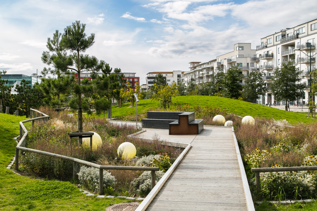 A scenic urban park featuring a wooden walkway, benches, and modern apartment buildings.
